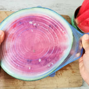 Person holding a watermelon with a colorful silicone lid on a wooden cutting board.