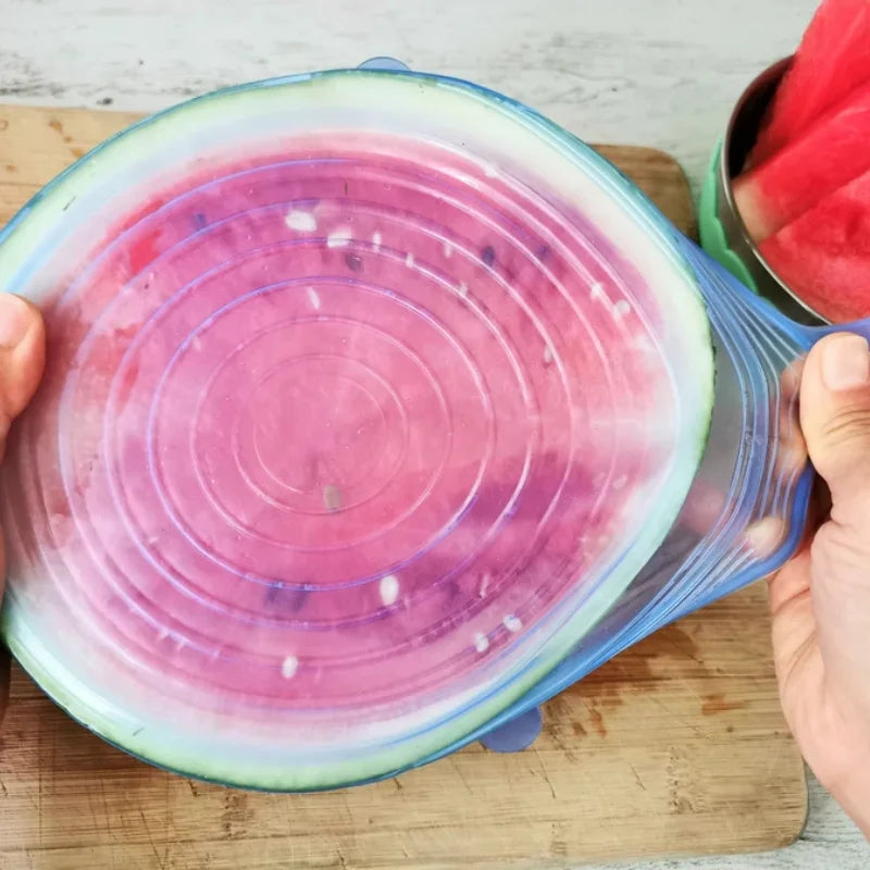 Person holding a watermelon with a colorful silicone lid on a wooden cutting board.