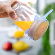 Clear container with orange juice and blue lid held by a hand, blurred background with fruits